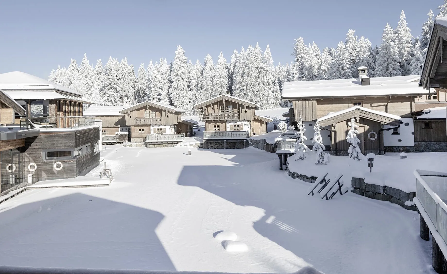 Verschneite Chalets des INNs HOLZ Chaletdorfs in einer Winterlandschaft mit schneebedeckten Bäumen im Hintergrund unter klarem Himmel