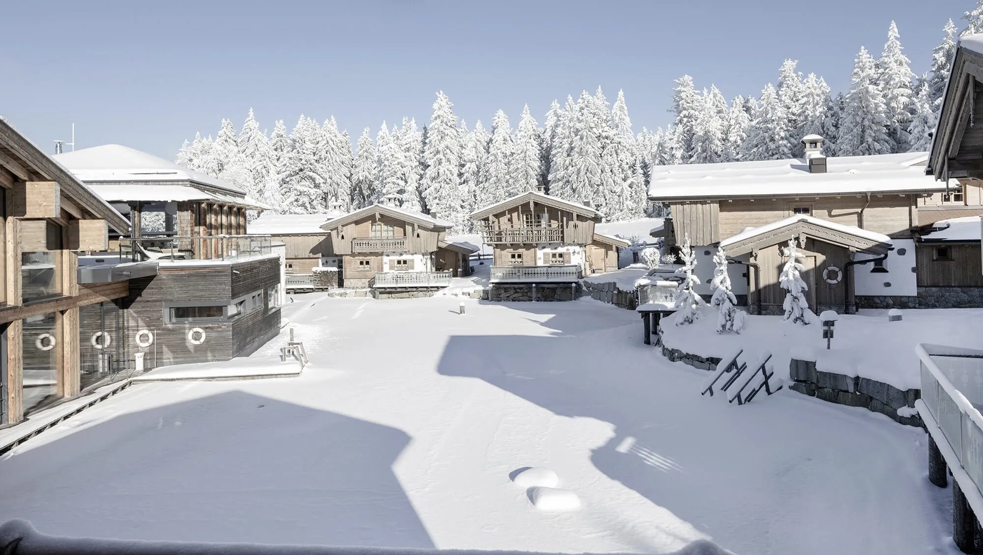 Verschneite Chalets des INNs HOLZ Chaletdorfs in einer Winterlandschaft mit schneebedeckten Bäumen im Hintergrund unter klarem Himmel