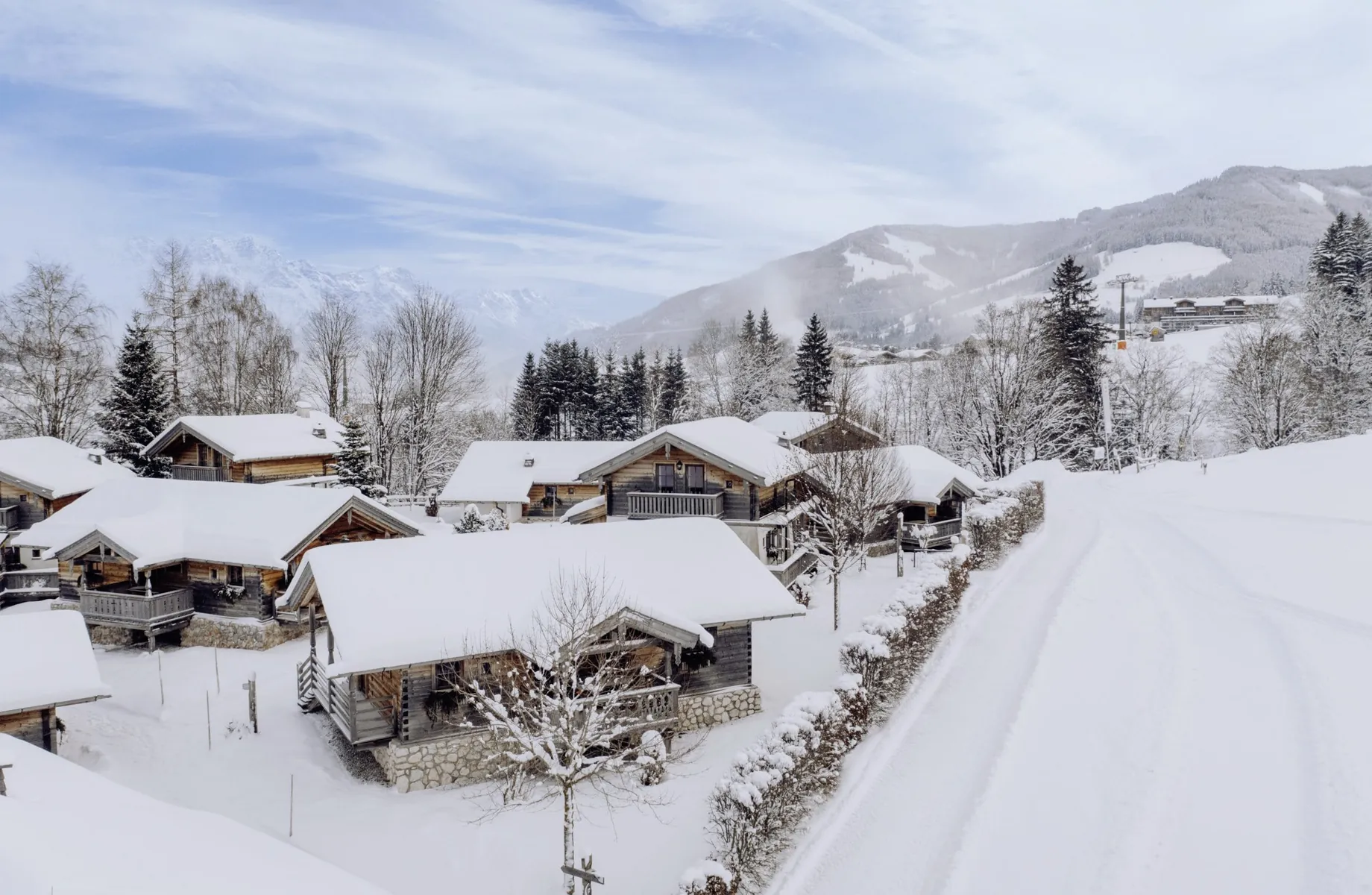 Verschneites Chaletdorf des Naturresort PURADIES mit schneebedeckten Dächern vor bewaldetem Berg