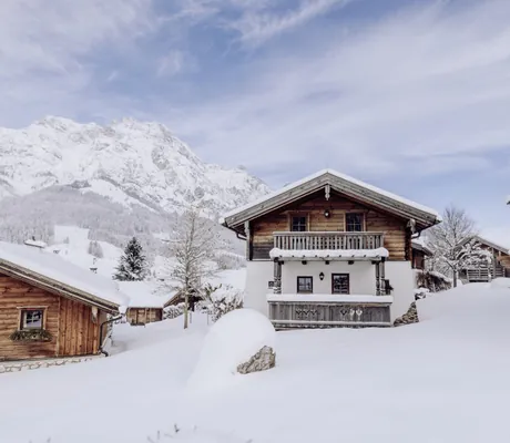 Verschneites Chaletdorf des Naturresort PURADIES vor schneebedeckten Bergen unter bewölktem Himmel