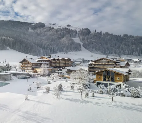 Verschneite Winterlandschaft und Blick auf das Naturresort PURADIES mit Outdoorpool vor bewaldeten Bergen unter bewölktem Himmel