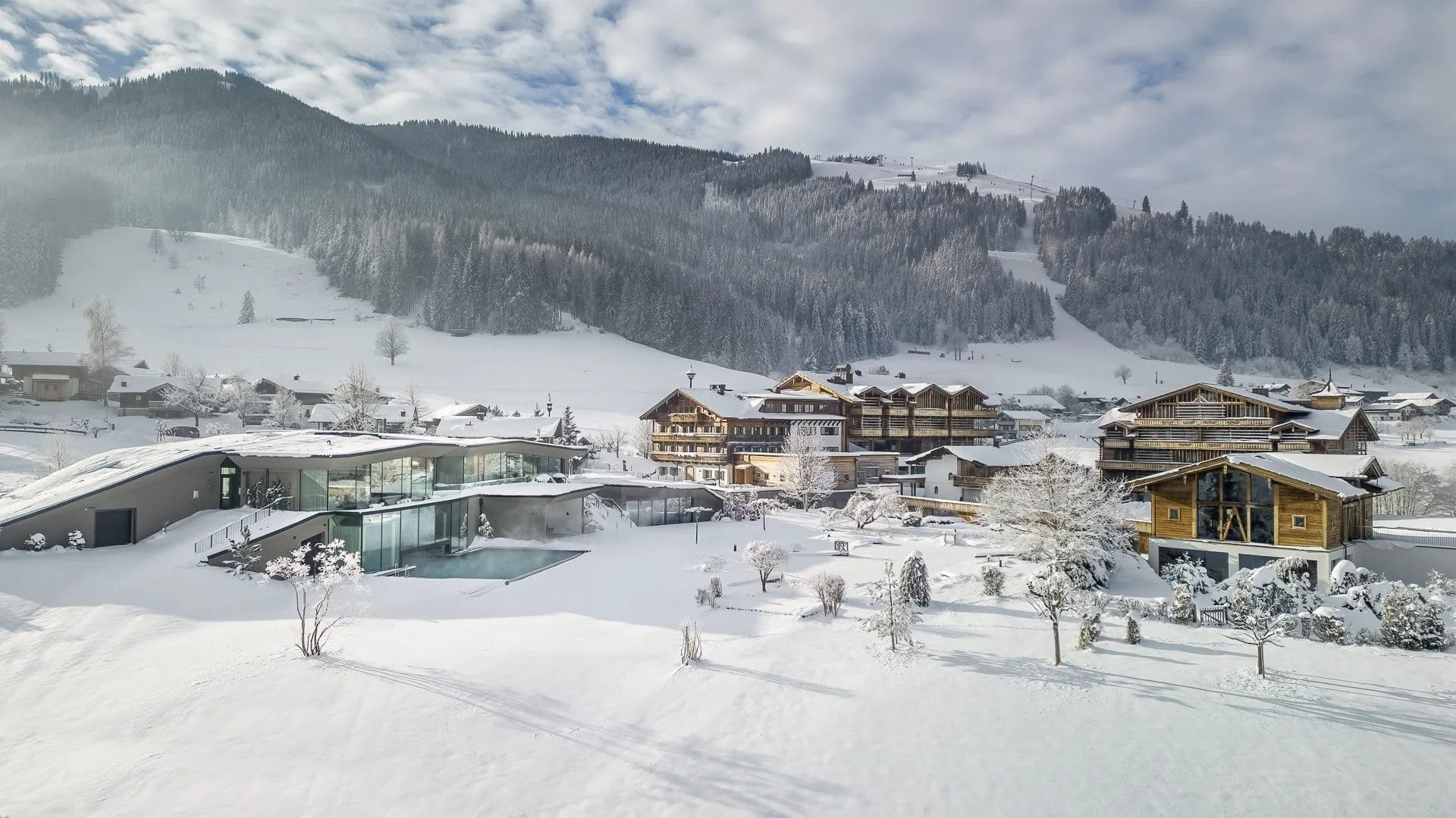 Verschneite Winterlandschaft und Blick auf das Naturresort PURADIES mit Outdoorpool vor bewaldeten Bergen unter bewölktem Himmel