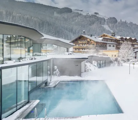 Outdoorpool des Naturresort PURADIES mit dampfendem Wasser vor verschneiter Hotelanlage und Bergen unter bewölktem Himmel