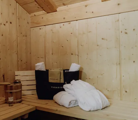Sauna eines Chalets im Naturresort mit Holzeimer und Kelle, zwei weißen Handtüchern und zwei weißen Bademänteln neben einer Tasche mit der Aufschrift 'PURADIES'