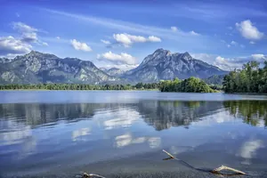 Berglandschaft mit bewaldeten Hügeln und Gipfeln, spiegelnd im ruhigen Forggensee unter blauem Himmel mit Wolken
