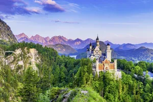 Schloss Neuschwanstein auf einem bewaldeten Hügel mit Alpen im Hintergrund unter blauem Himmel mit Wolken