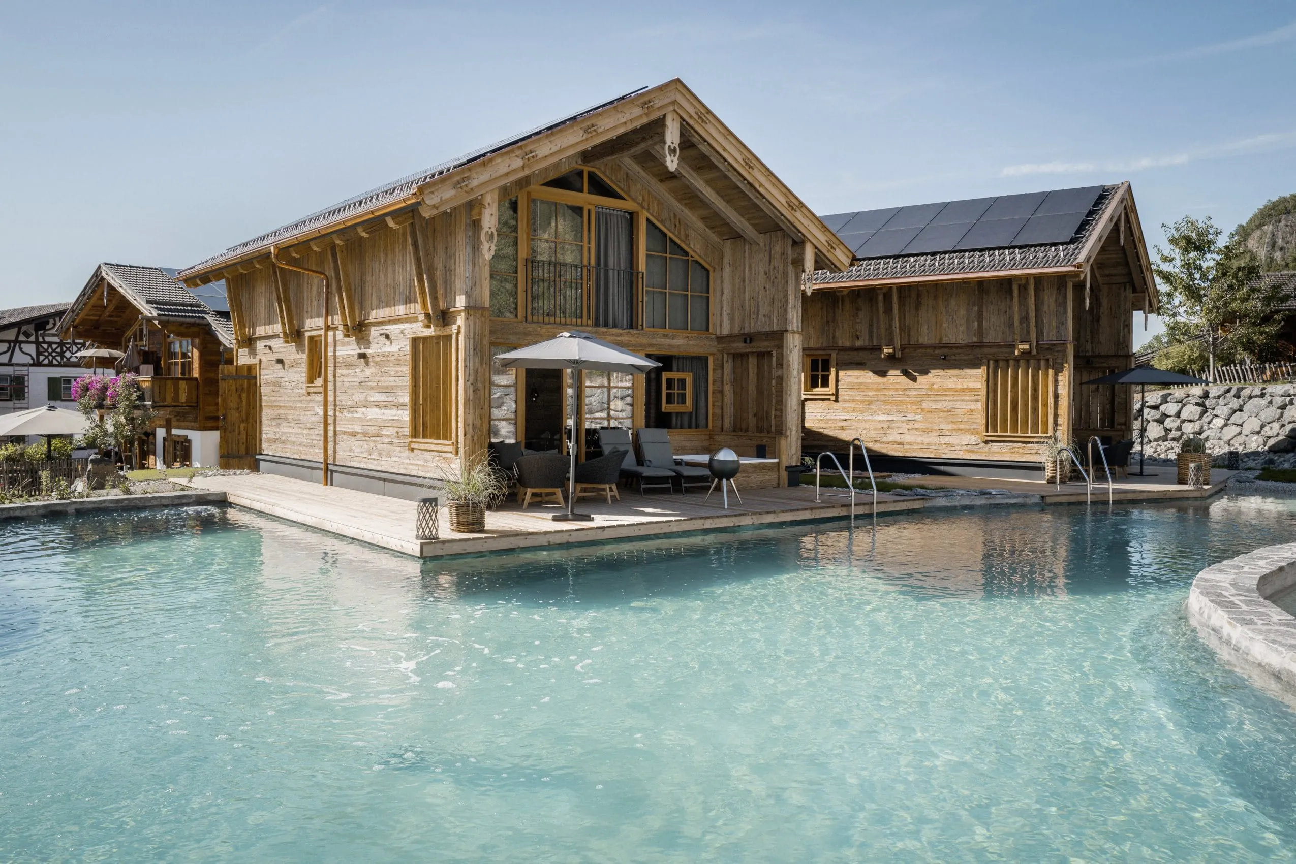 Neues Chalet der Alpzitt-Chalets mit Terrasse und Sonnenschirm direkt am klaren Naturbadesee unter blauem Himmel