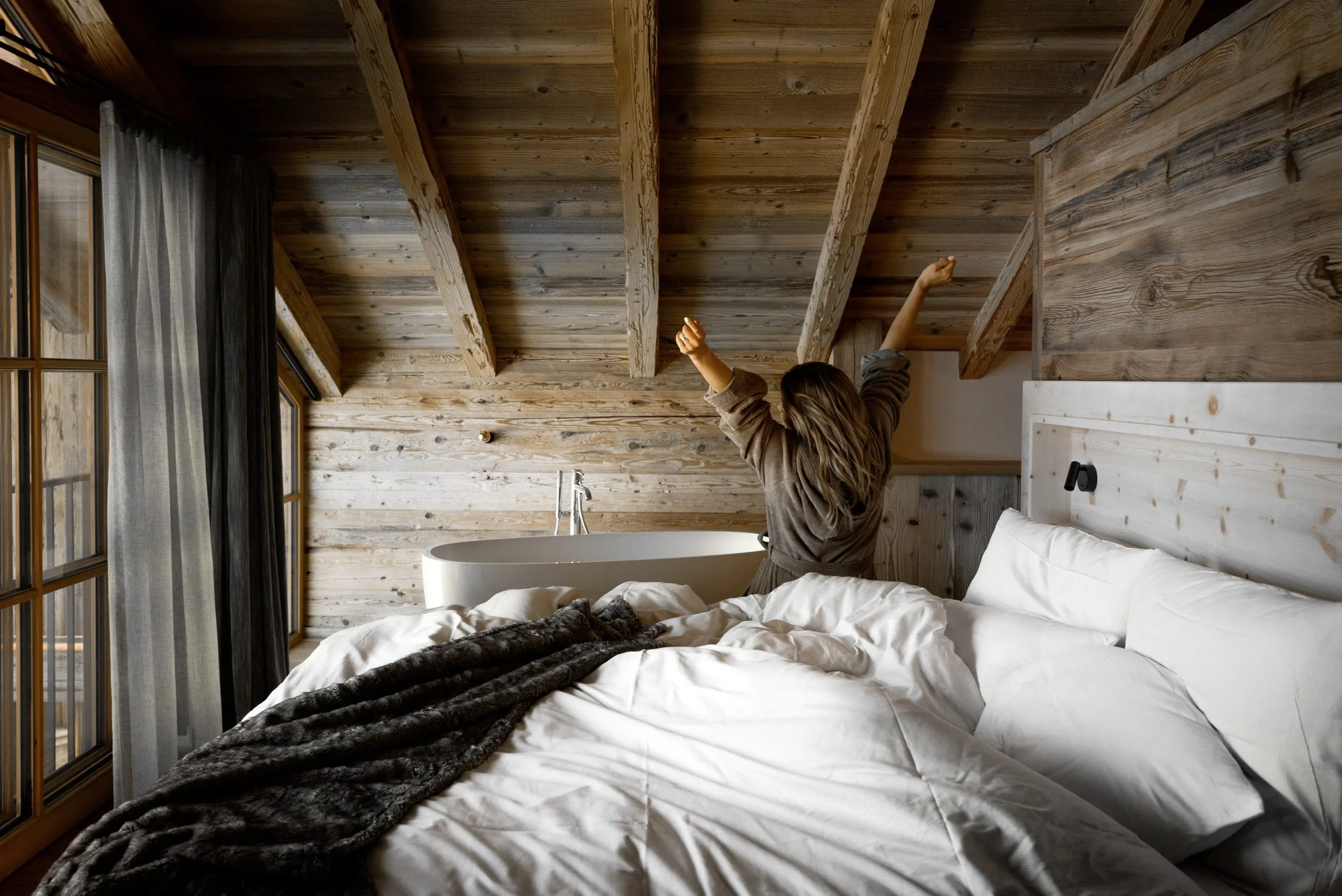 Frau mit langen Haaren und Bademantel sitzt auf einem Bett der Alpzitt-Chalets und streckt die Arme, freistehende Badewanne im Hintergrund