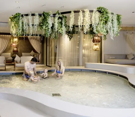 Familie mit zwei kleinen Kindern sitzt in rund geschwungenem Kinderpool mit Wasserrutsche, darüber hängende grüne Pflanzen, im Hintergrund gemütliche Liegen