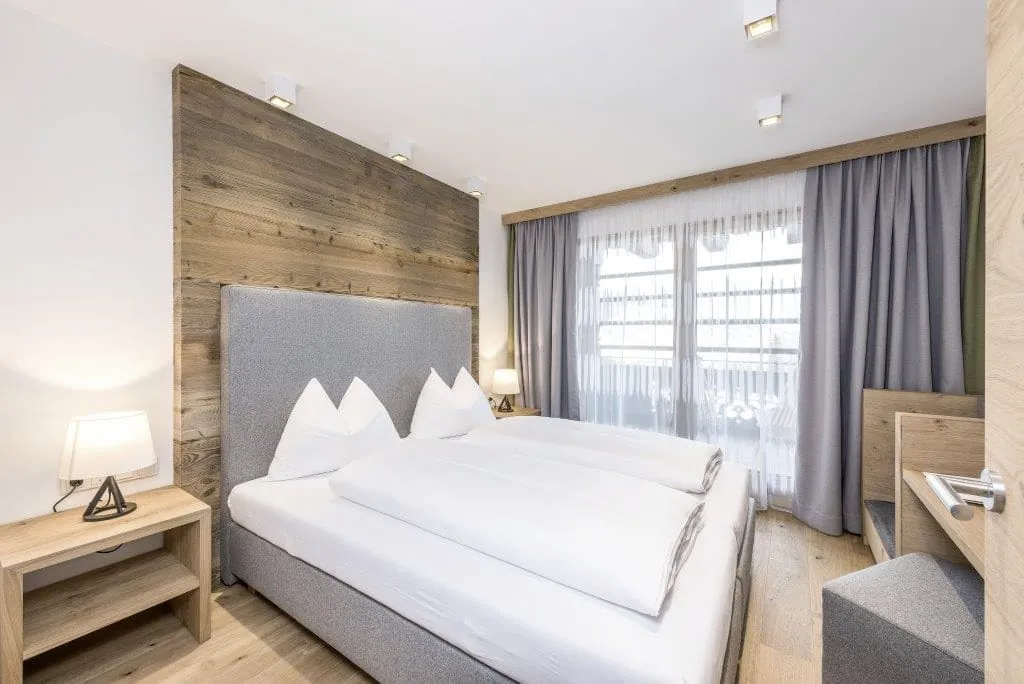 Modern eingerichtetes Hotelzimmer im Naturresort PURADIES mit Doppelbett, grauem Polsterkopfteil, Holzwand, Nachttisch mit Lampe und großem Fenster mit grauen Vorhängen