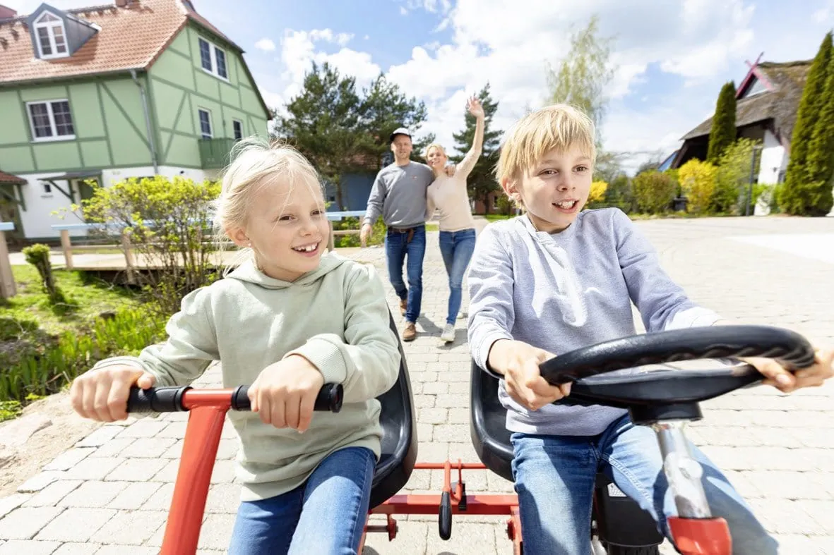 Zwei Kinder fahren mit Tretfahrzeugen auf dem Gel&auml;nde des BEECH Resort Fleesensee, Eltern winken im Hintergrund