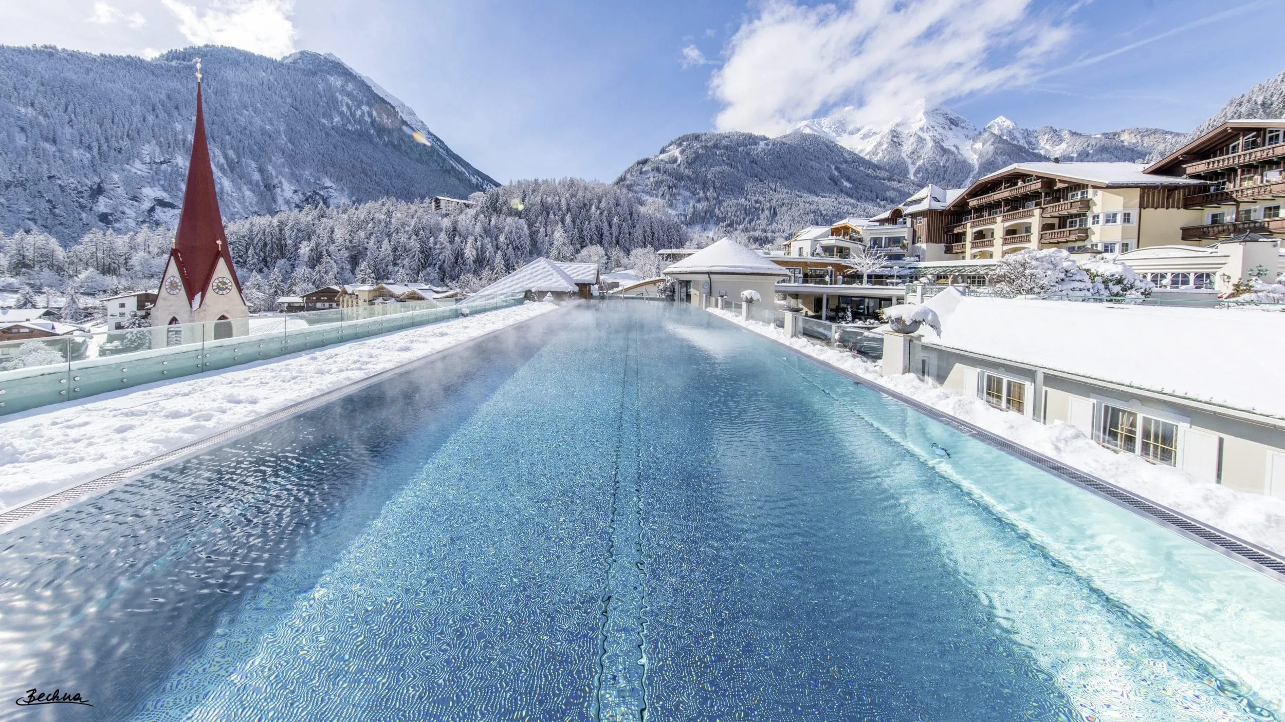 Langer Outdoorpool auf dem Hoteldach des STOCK resort, umgeben von schneebedeckten Bergen und Geb&auml;uden und einem Kirchturm auf der linken Seite