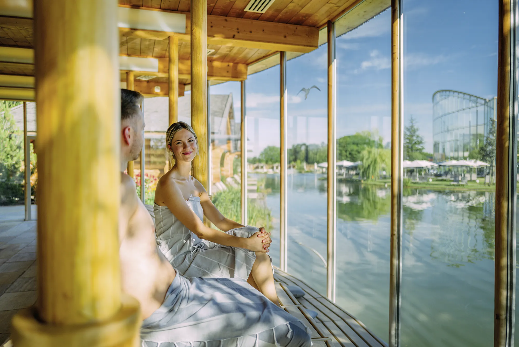 P&auml;rchen sitzt mit Saunat&uuml;chern in der Seesauna der Therme Erding, durch bodentiefe Fenster blicken sie auf den umgebenden See