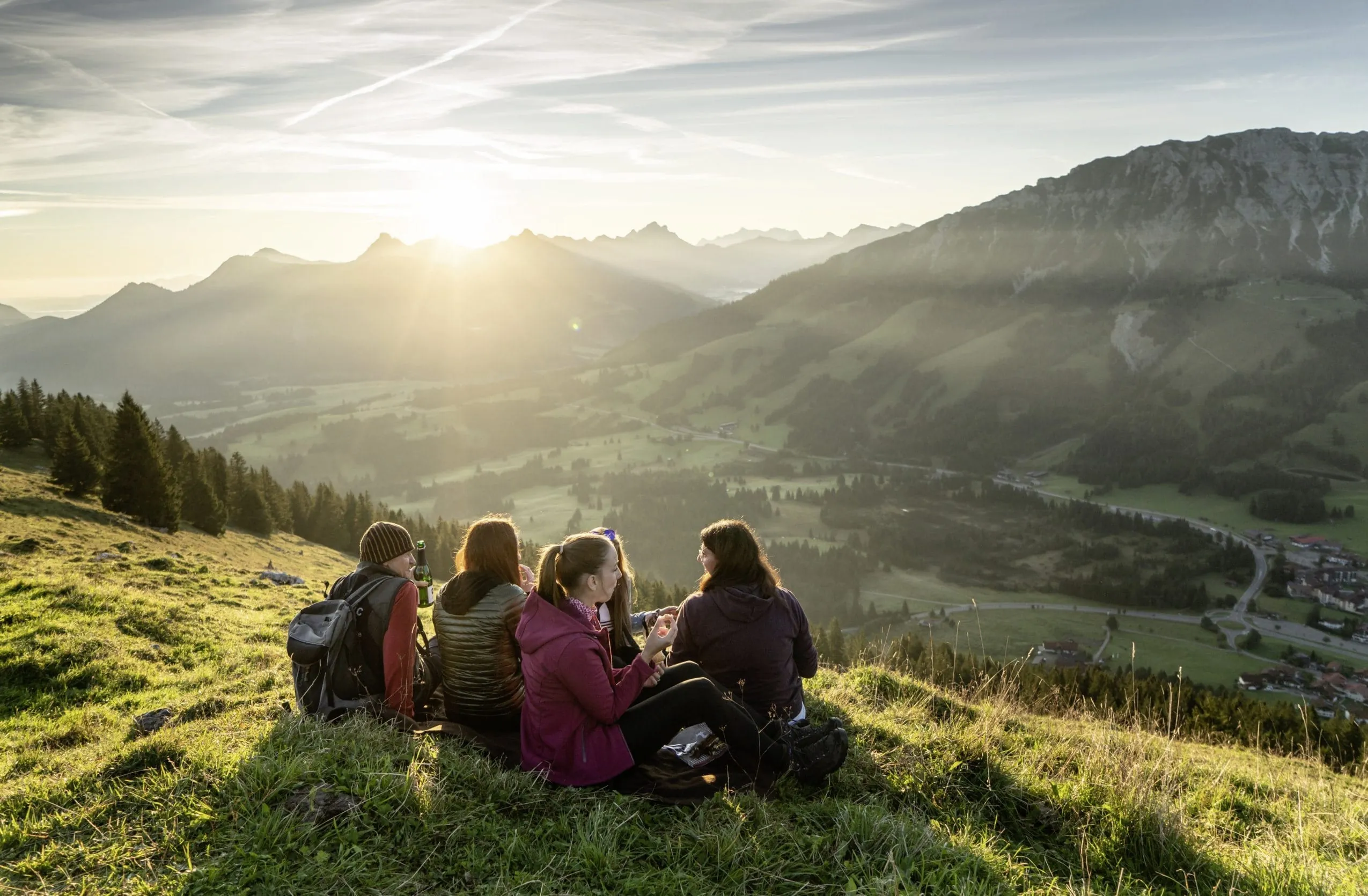 Gruppe junger Menschen sitzt zusammen mit dem Bergf&uuml;hrer auf einem Allg&auml;uer Bergkamm und blickt Richtung Sonnenuntergang