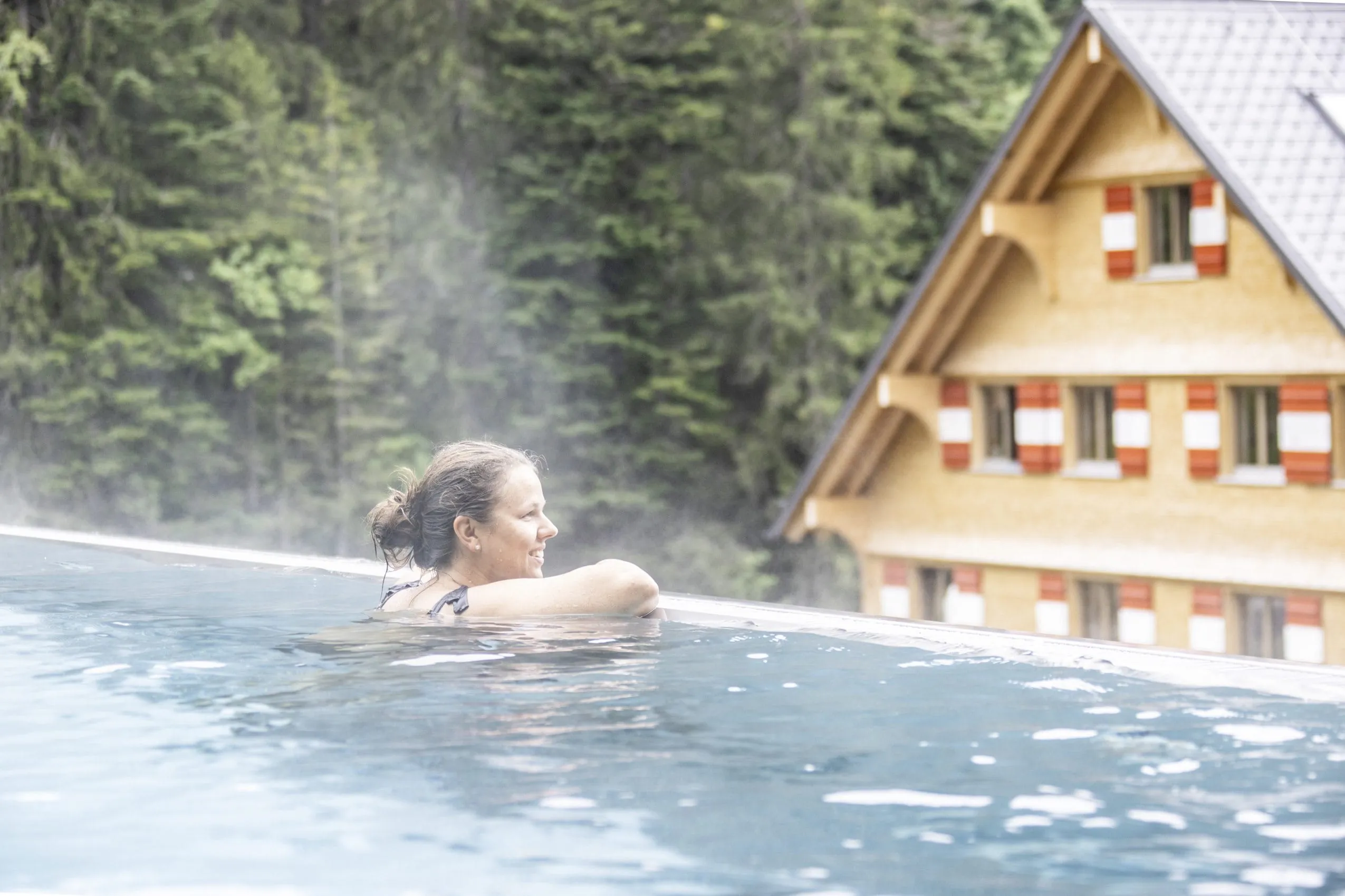 Frau entspannt im Outdoorpool des Berghaus Schr&ouml;cken mit Bergpanorama und Wanderhotel im Hintergrund