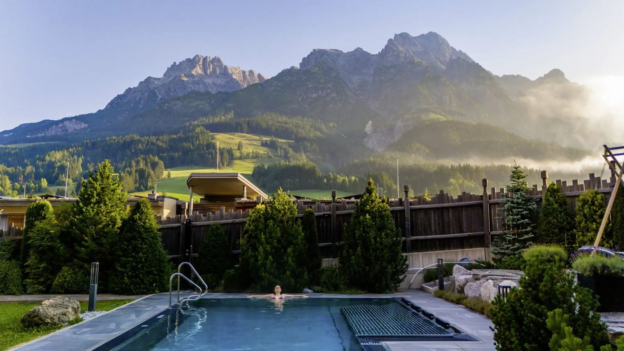 Frau entspannt im Outdoorpool des Wanderhotel Good Life Resort Riederalm in Leogang, im Hintergrund Bergpanorama im Morgenlicht