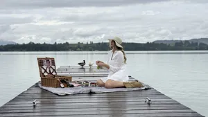 Frau sitzt mit weißem Kleid und Sonnenhut auf einem Steg am See und macht Picknick