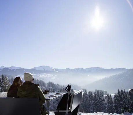 Paar genießt Bergpanorama mit Sonnenschein auf Aussichtsplattform in verschneiter Winterlandschaft bei Oberstaufen