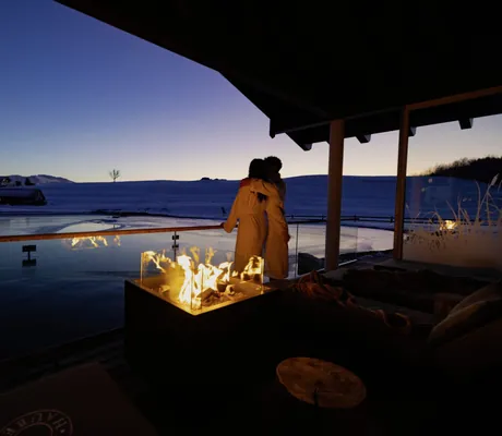 Paar an beleuchteter Feuerschale auf Terrasse mit Blick auf verschneite Landschaft in der Abenddämmerung