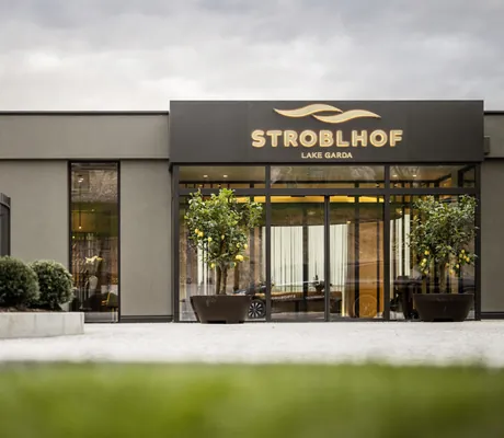 Eingang des Wellnesshotels STROBLHOF LAKE GARDA Active Family SPA Resort mit Glasfront, Pflanzen und schwarzem Schild mit goldener Aufschrift