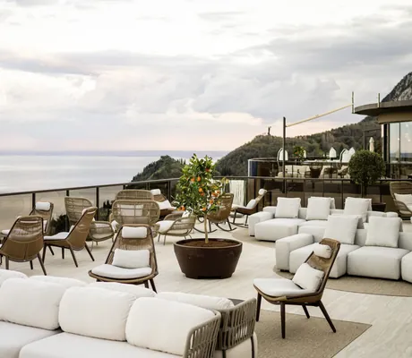 Rooftop-Terrasse des STROBLHOF LAKE GARDA Active Family SPA Resort mit weißen Polstersofas, Rattanstühlen, Orangenbaum, Blick auf See und Berge unter bewölktem Himmel