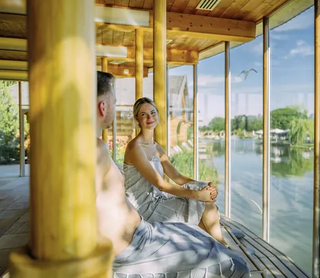 Junges Paar mit Handtüchern umgewickelt sitzt in einer Sauna des Hotel Victory Therme Erding mit großen Fenstern und Blick auf einen See