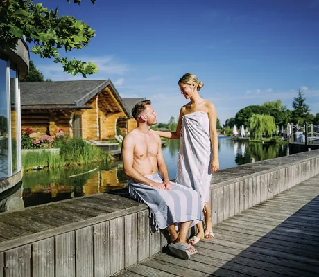 Mann mit Handtuch um die Hüfte sitzt auf Holzsteg, Frau mit Handtuch steht neben ihm, im Hintergrund Holzhaus und See des Hotel Victory Therme Erding