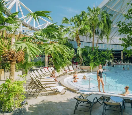 Innenpool mit mehreren Personen, umgeben von Liegestühlen und Palmen unter einer halboffenen Glaskuppel im Hotel Victory Therme Erding