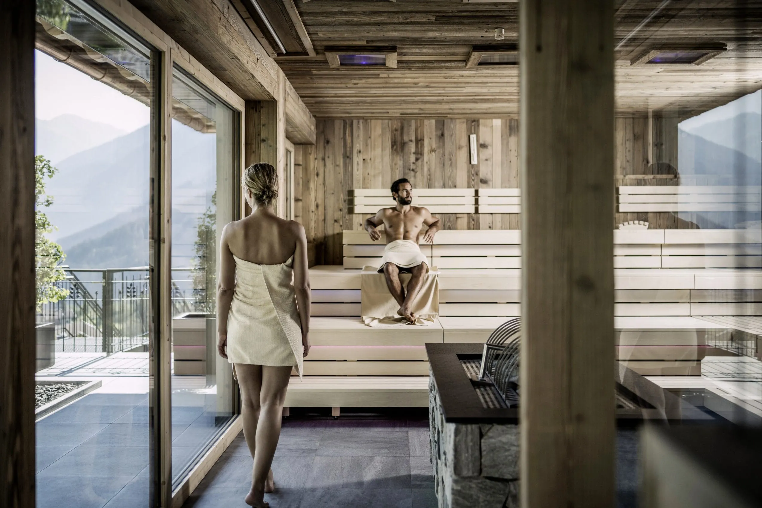 Frau mit wei&szlig;em Handtuch steht in einer Sauna im STOCK resort, Mann sitzt auf Holzbank, gro&szlig;e Fenster mit Bergblick