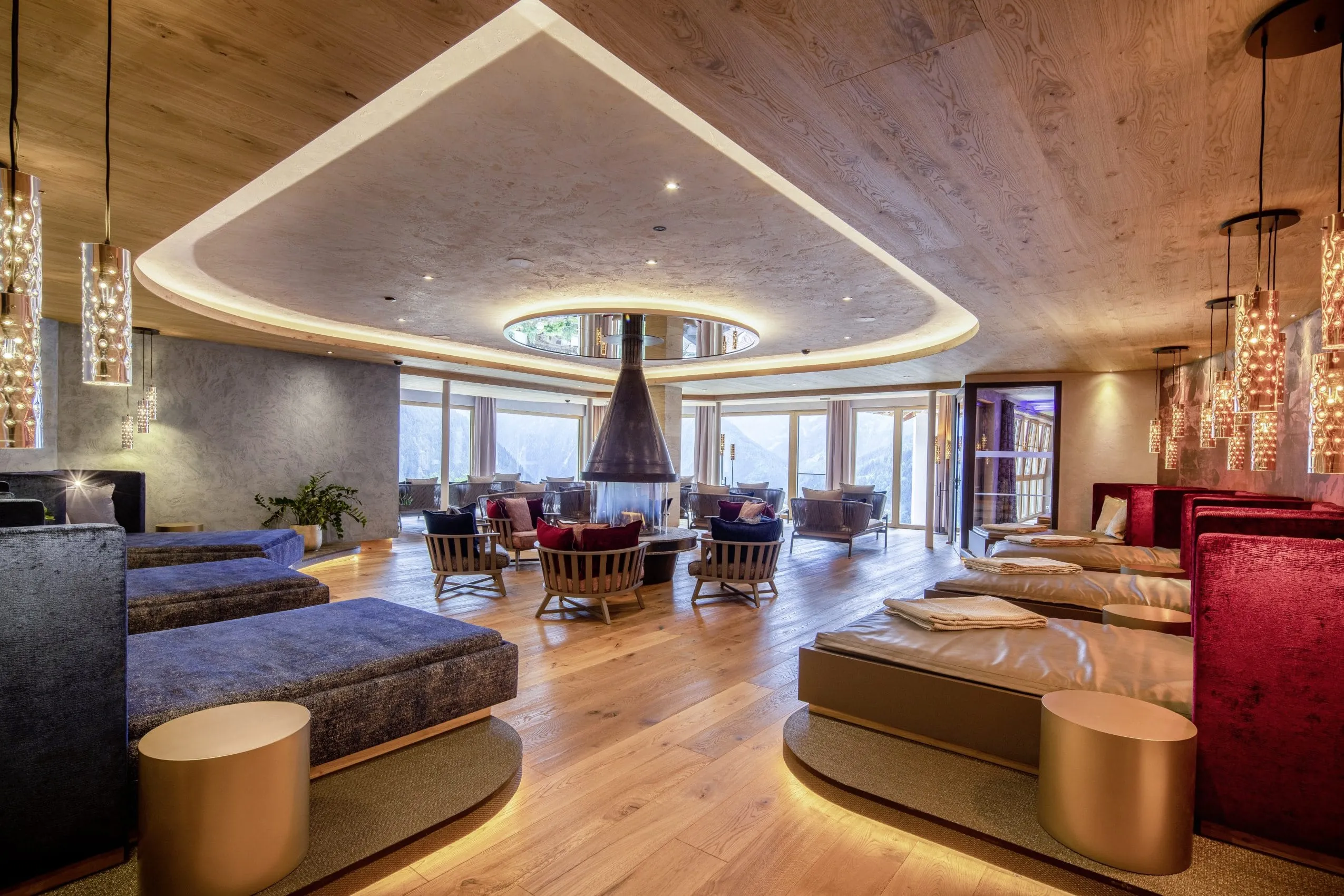 Gro&szlig;er, moderner Panoramaruheraum im STOCK resort mit Holzdecke und -boden, zentralem Kamin, Sessel und Liegen, gro&szlig;en Fenstern mit Bergblick