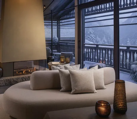 Gemütlicher Innenraum eines Chalets mit modernem Kamin, Sofa und Panoramablick auf die verschneite Winterlandschaft