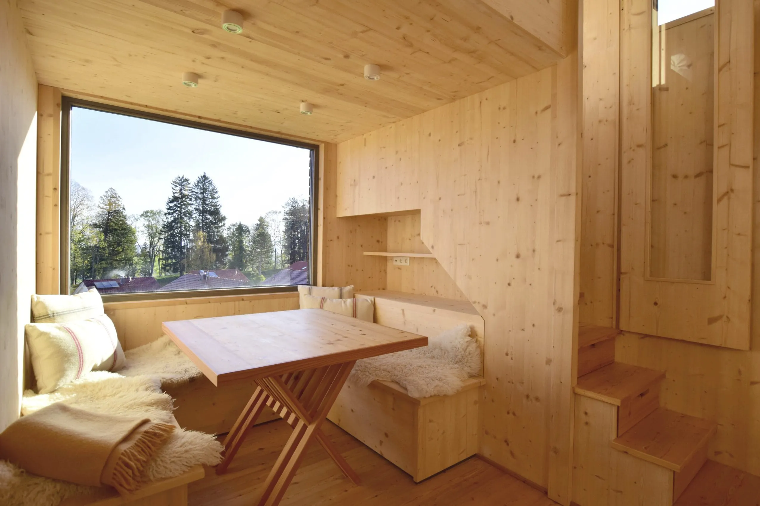 Gemütliche Sitzecke mit Holztisch und mit Fell belegten Bänken vor einem großen Panoramafenster in einem Tiny House des SEINZ Wisdom Resorts, mit Blick in die Natur