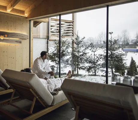Mann in weißem Bademantel sitzt am Panoramafenster des Ruheraums in Kesslers Mountain Lodge und schaut in Schneelandschaft