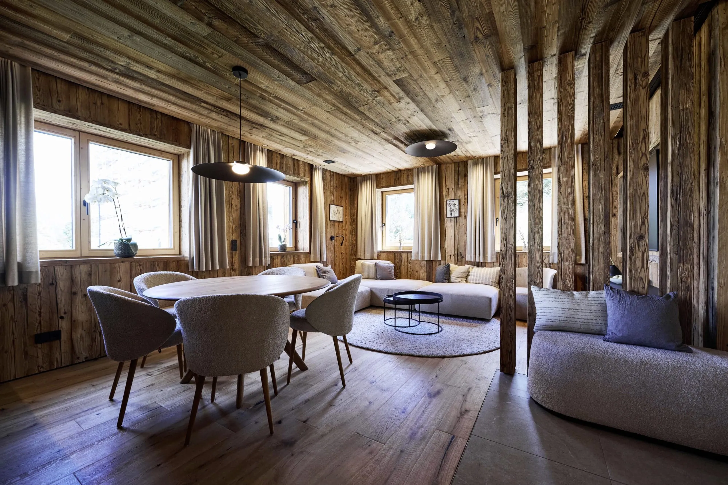 Wohn- und Essbereich in den Ziegelberg Chalets mit Holzw&auml;nden und -decke, runder Esstisch mit gepolsterten St&uuml;hlen, Sofa und Couchtisch auf rundem Teppich
