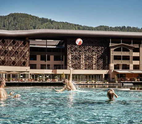 Drei Personen spielen mit einem rot-weißen Wasserball im Außenpool vor dem großen Hotelgebäude des Falkensteiner Family Resort Lido