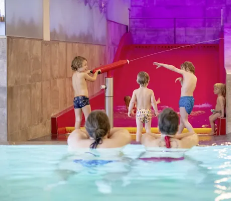 Mehrere Kinder spielen an einer roten Wasserrutsche, zwei Erwachsene beobachten sie vom Wasser aus