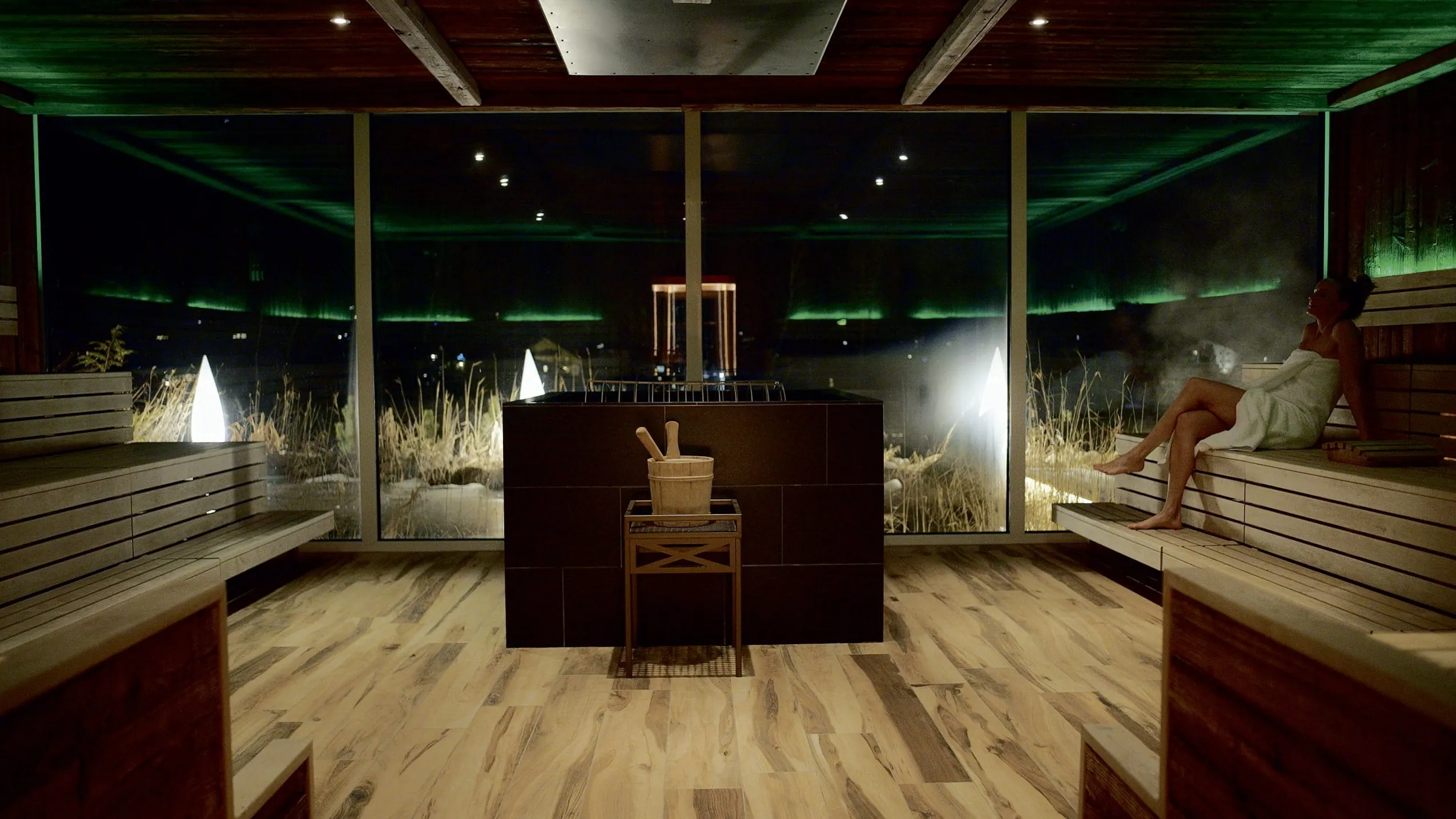 Frau sitzt in stilvoller Sauna im Alpin Resort Sacher Seefeld mit gro&szlig;em Fensterblick, zentralem Saunaofen und entspannter Atmosph&auml;re bei Nacht
