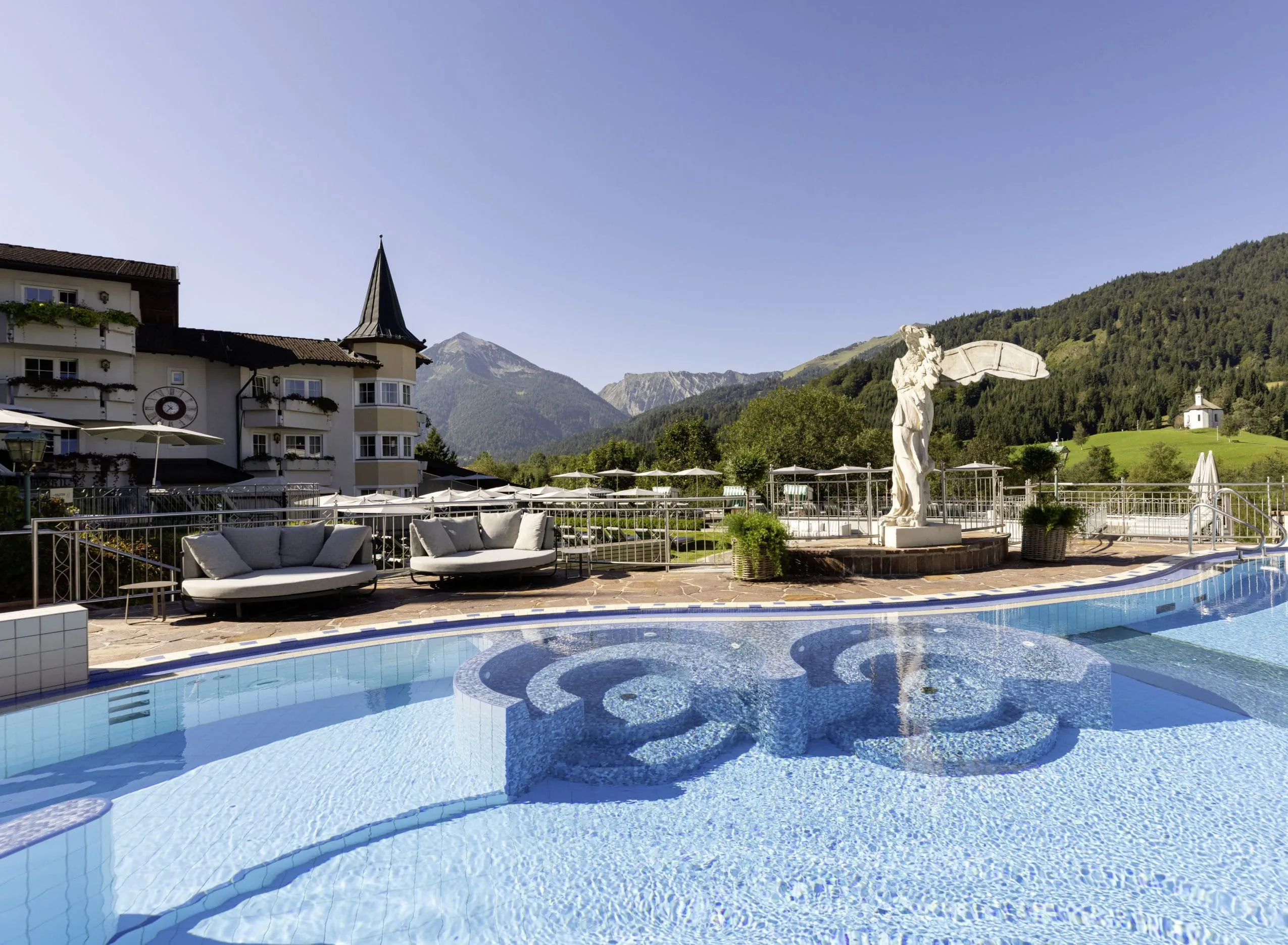 Au&szlig;enpool des Posthotel Achenkirch mit Statue, gem&uuml;tlichen Liegen, Hotelfassade und Berglandschaft im Hintergrund