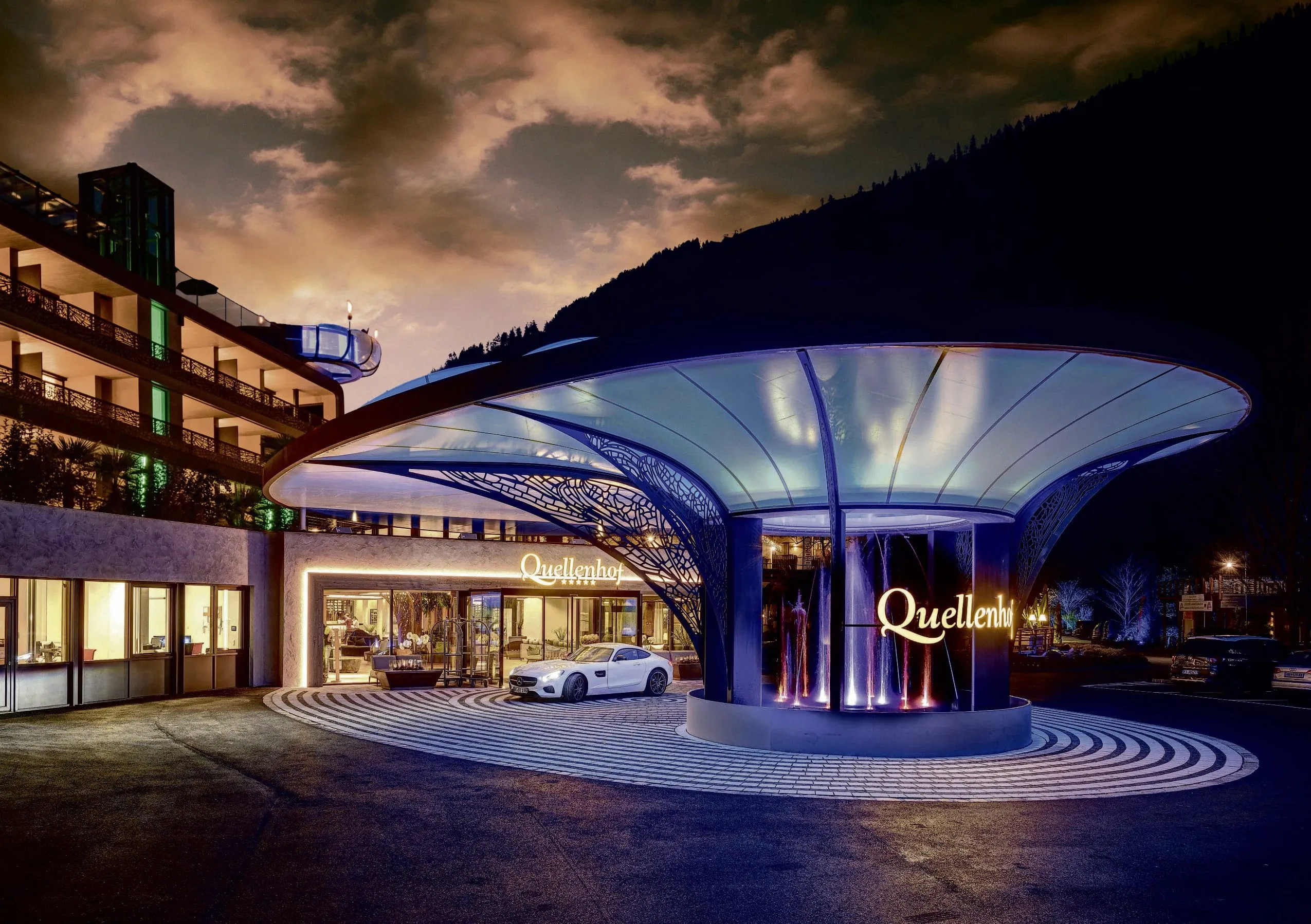 Beleuchteter, luxuri&ouml;ser Eingangsbereich des Quellenhof Luxury Resort Passeier mit moderner &Uuml;berdachung und Wasserspiel bei Abendstimmung