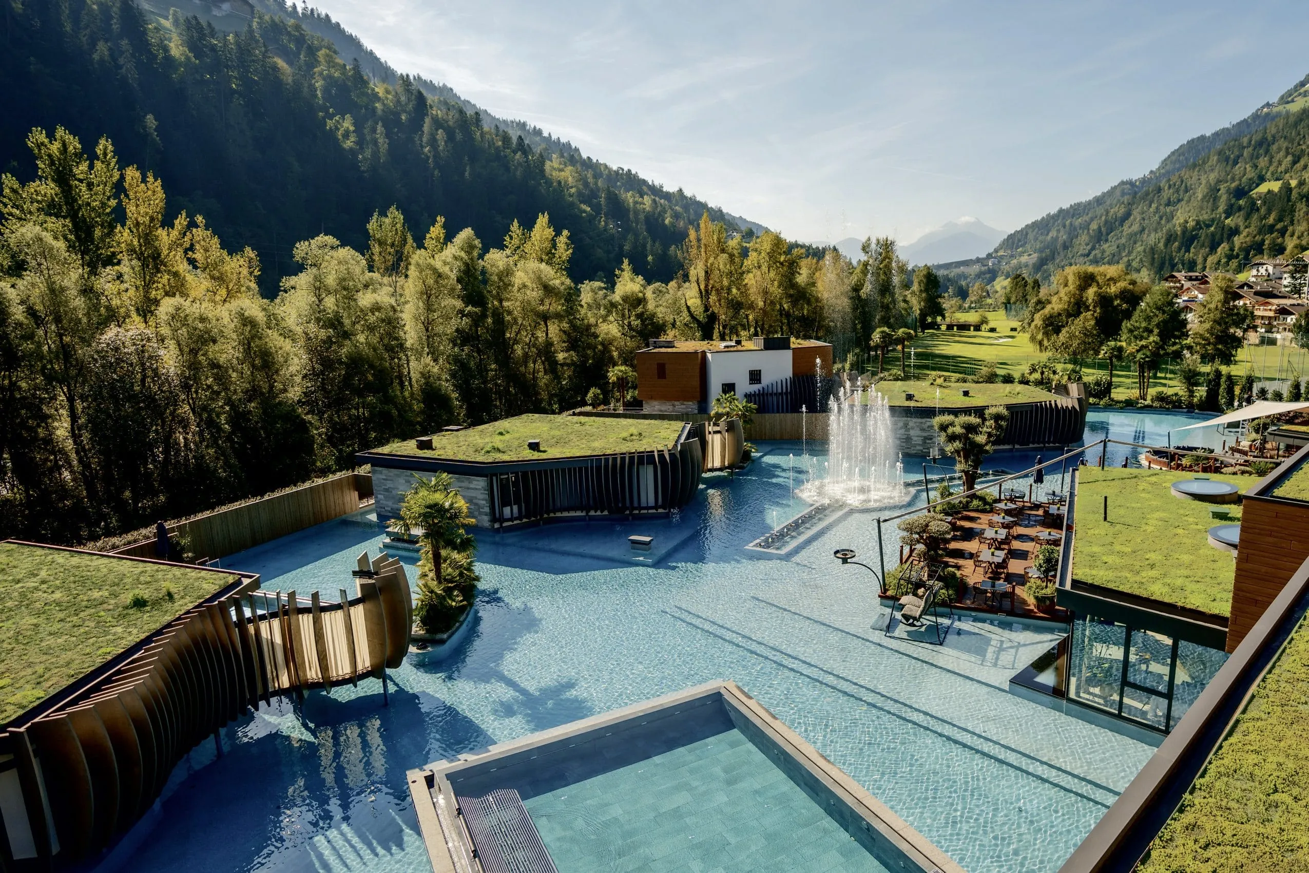 Blick auf die gro&szlig;e Poolanlage der Quellenhof See Lodge mit Springbrunnen, gr&uuml;nen D&auml;chern und m&ouml;blierter Holzterrasse