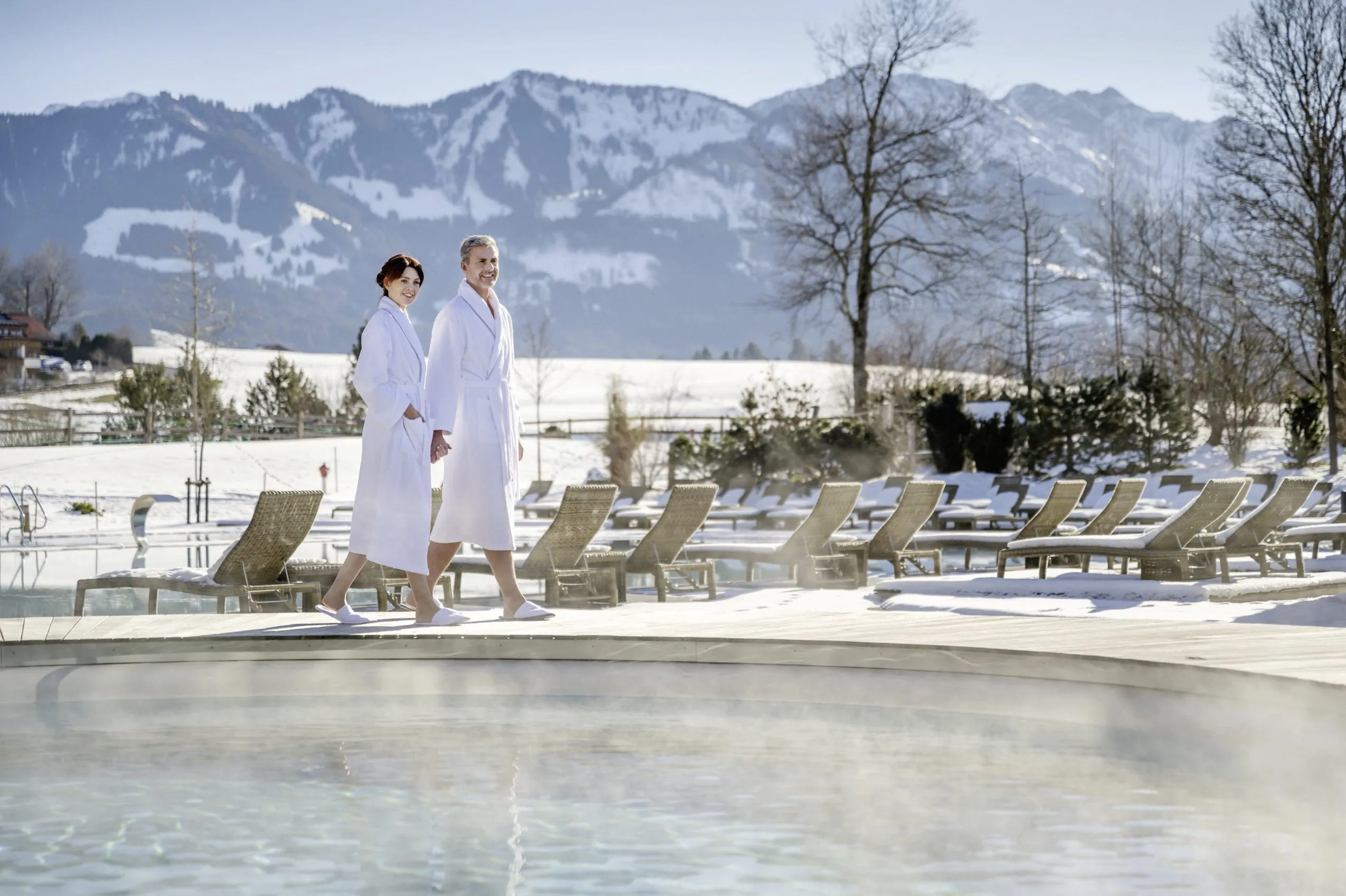 Paar im Bademantel am Outdoorpool des Sonnenalp Resorts im Winter, mit Blick auf schneebedeckte Berge