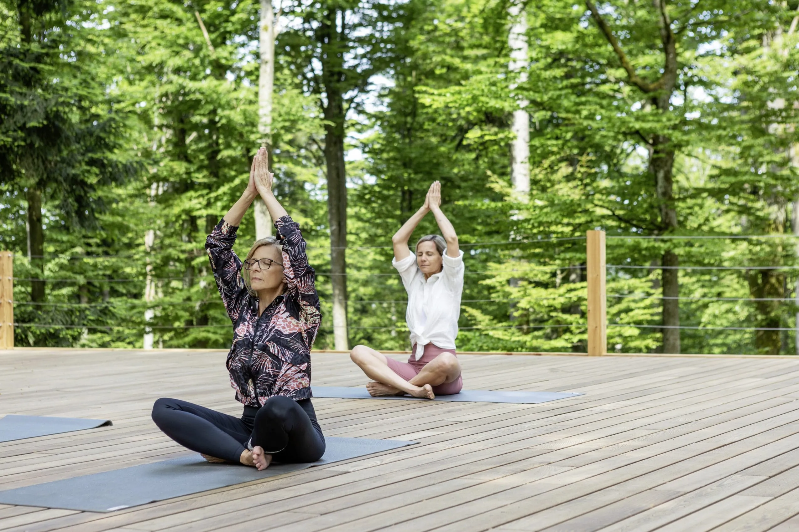 Zwei Frauen beim Yoga auf einer h&ouml;lzernen Plattform im Wald Spa des Wellness- und Naturresort Reischlhof, umgeben von gr&uuml;nem Laubwald