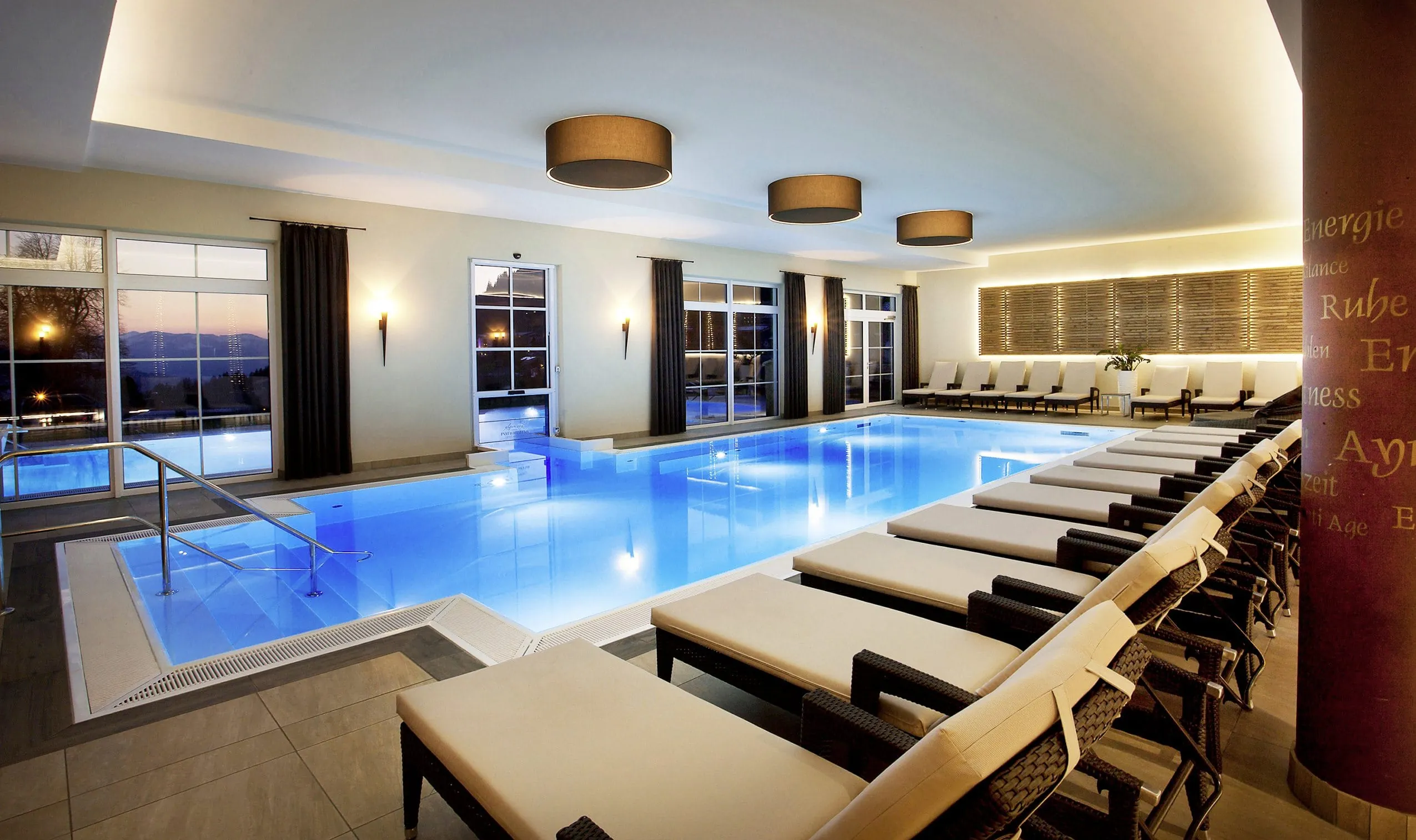 Beleuchteter Indoorpool im Panoramahotel Oberjoch mit Liegest&uuml;hlen, gro&szlig;en Fenstern und stilvoller Beleuchtung