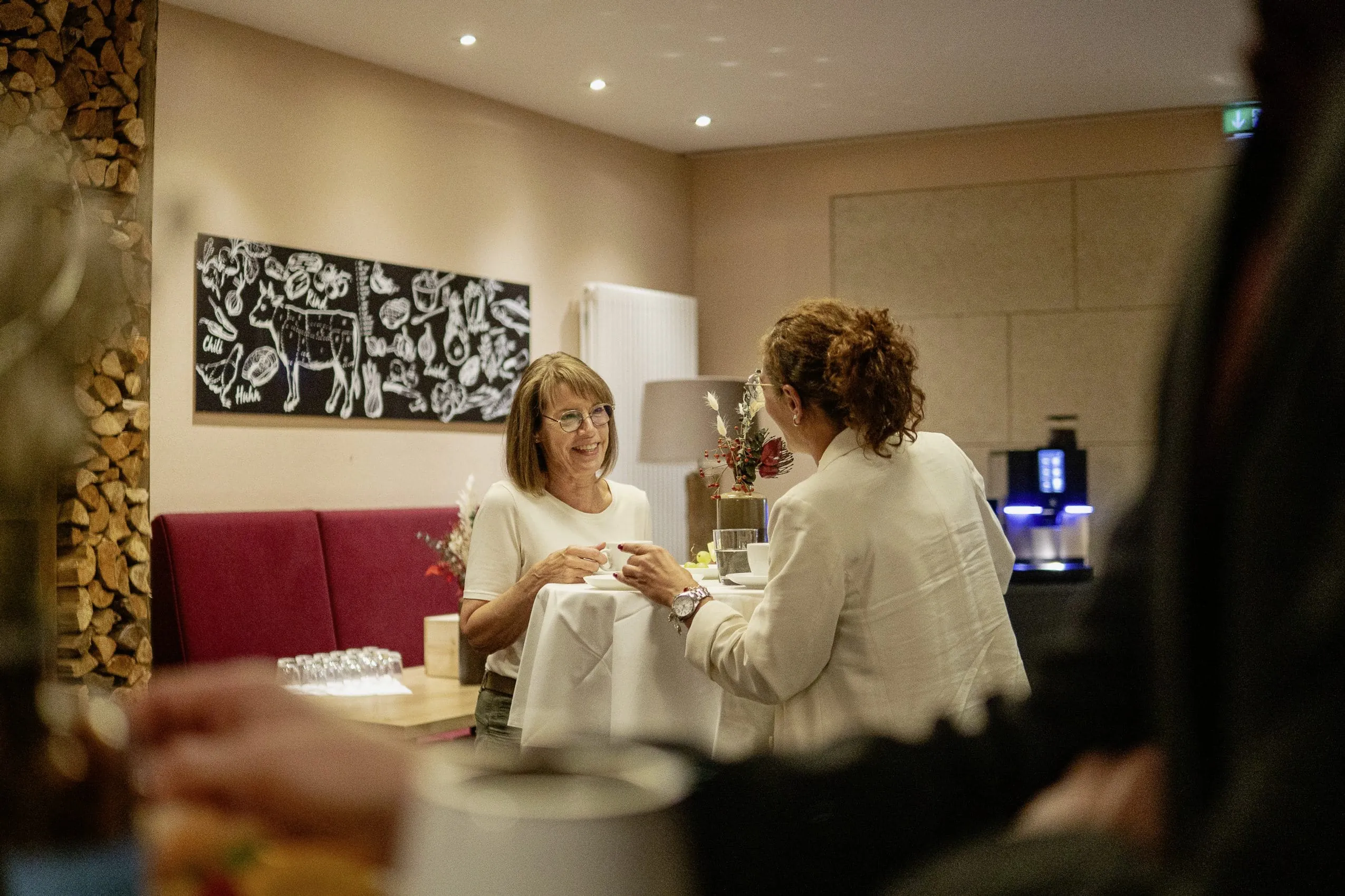 Zwei Frauen bei einer Kaffeepause an einem Stehtisch im Innenbereich des Panoramahotels Oberjoch, dekoriert mit Holz und Blumen