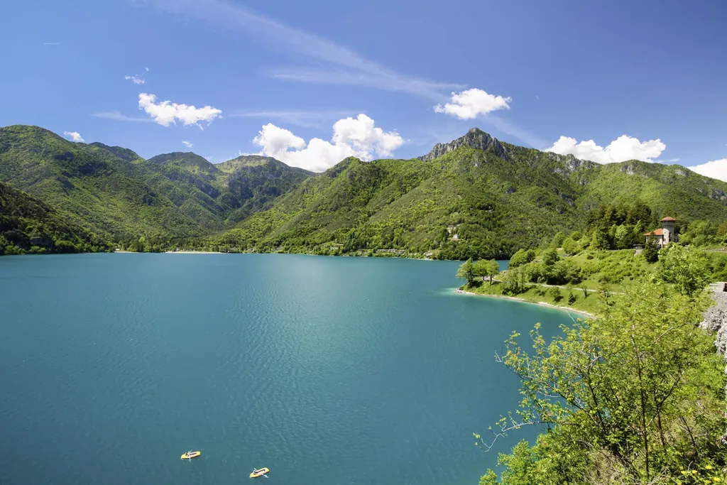 Blick auf den türkisblauen Ledrosee, umgeben von grünen Bergen unter klarem Himmel, mit zwei Schlauchbooten auf dem Wasser und einer Villa am Uferstreifen