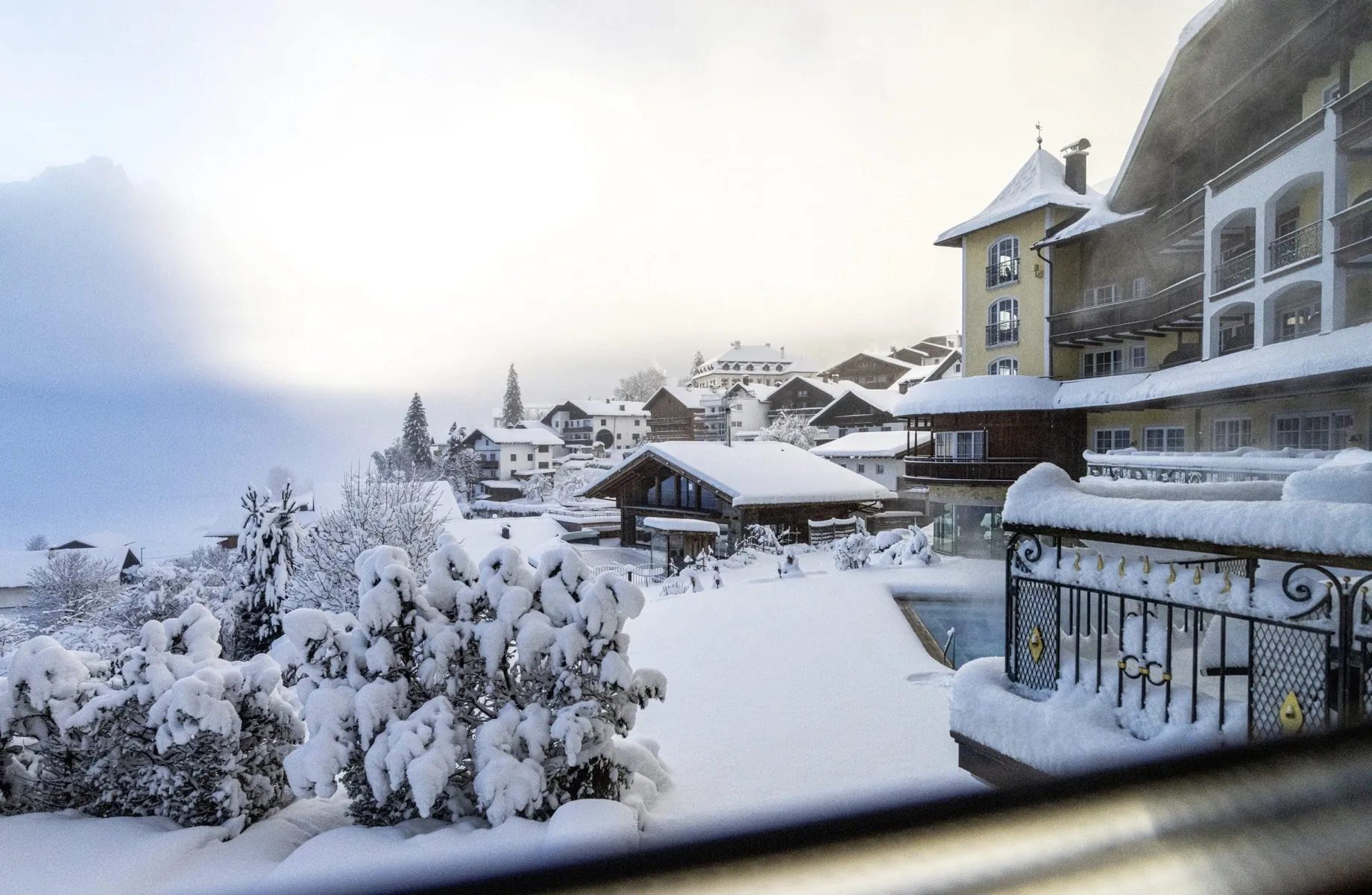 Fassade des Hotel Post Lermoos mit gelbem Turm im tiefen Schnee und Blick auf Zugspitze im Nebel