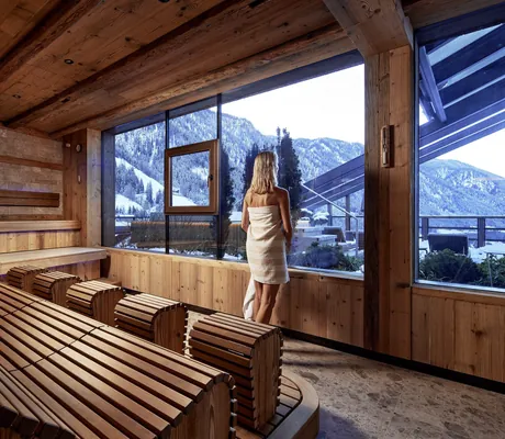 Frau mit Handtuch steht in einer Holzsauna des Hotel Quelle Nature Spa Resort und blickt auf verschneite Berge