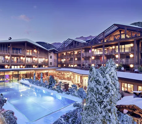 Hotelfassade des Hotel Quelle Nature Spa Resort mit beleuchtetem Außenpool und schneebedeckten Bäumen im Winter, umgeben von Bergen im Abendlicht