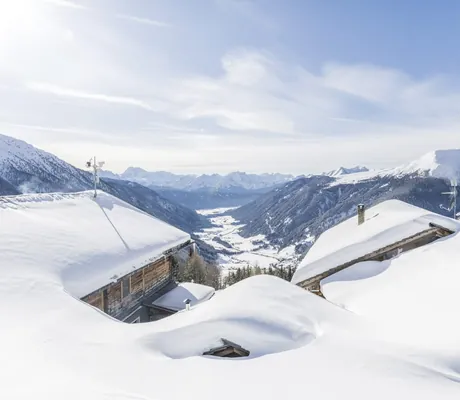 Verschneite Dächer von Holzhäusern in der Nähe des Hotel Quelle Nature Spa Resort vor einem Tal mit Bergen unter blauem Himmel
