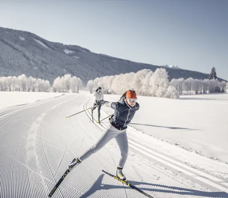Zwei Personen beim Langlaufen auf einer präparierten Loipe in einer verschneiten Winterlandschaft mit Bergen und Bäumen im Hintergrund in der Nähe des Hotel Quelle Nature Spa Resort